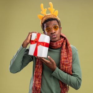 african-american-man-with-funny-rim-with-deer-horns-scarf-around-neck-holding-christmas-present-looking-camera-confused-standing-green-background_141793-39745